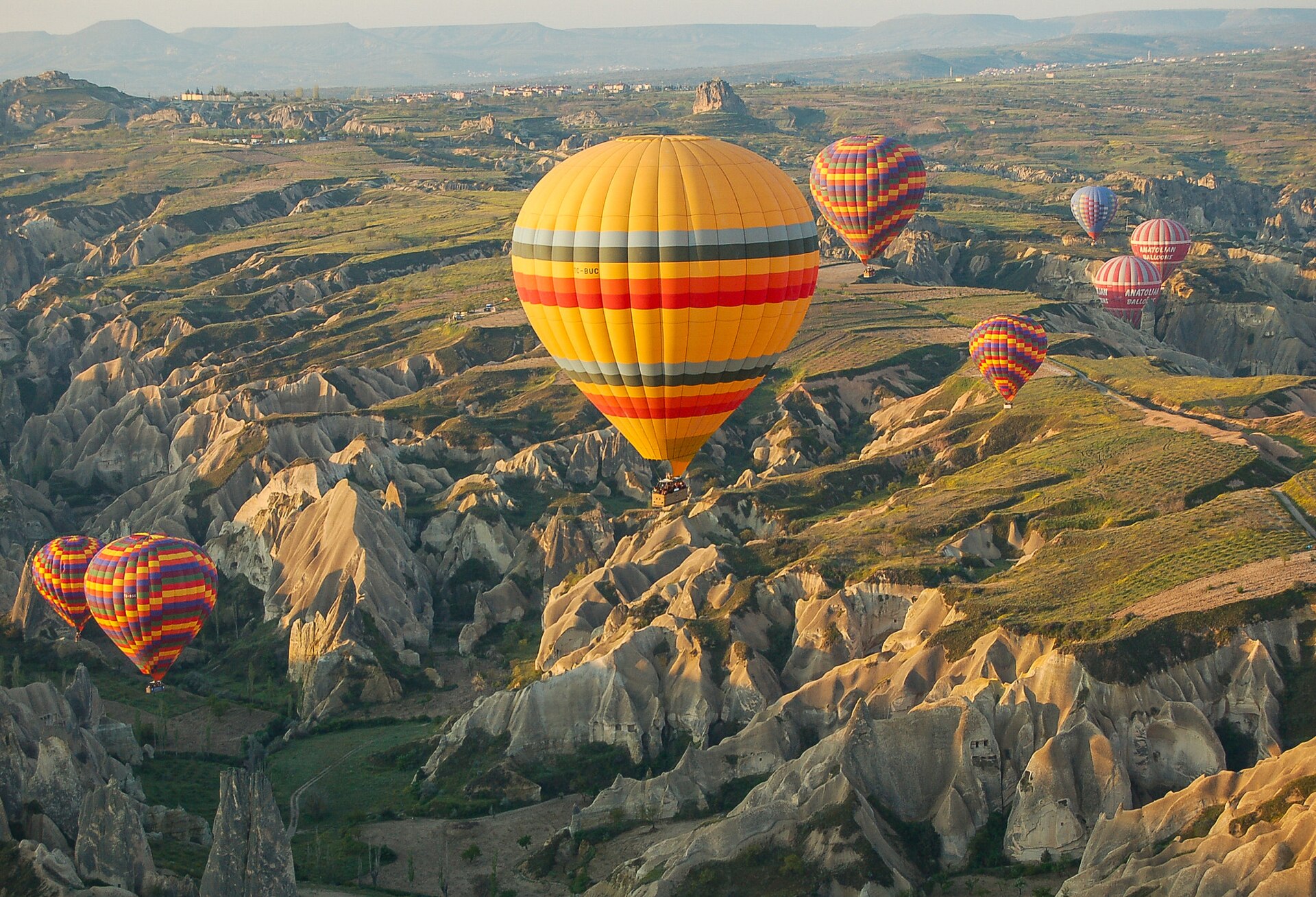 Mongolfiere colorate sorvolano le valli e le formazioni rocciose della Cappadocia, con un grande pallone giallo al centro.