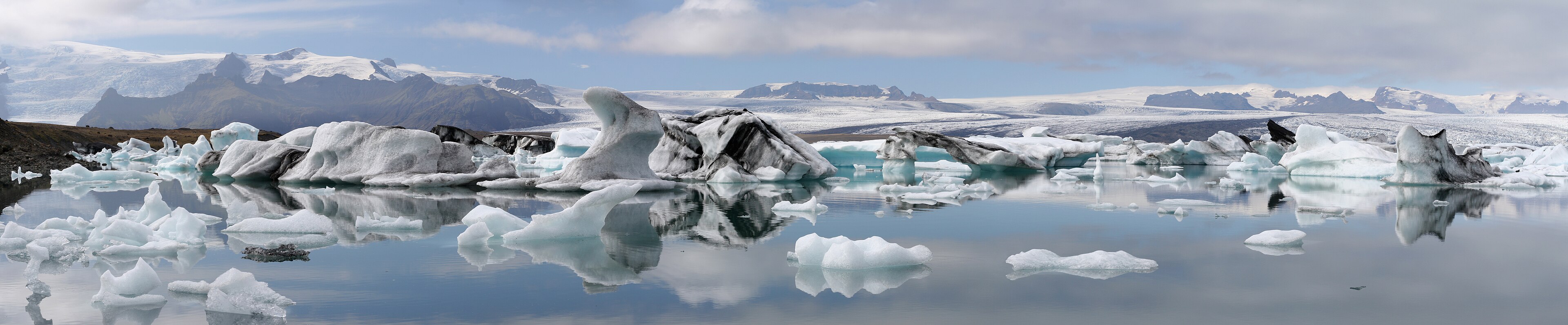 Iceberg bianchi e azzurri si riflettono nella laguna glaciale Jökulsárlón, con montagne innevate sullo sfondo, in pieno giorno.