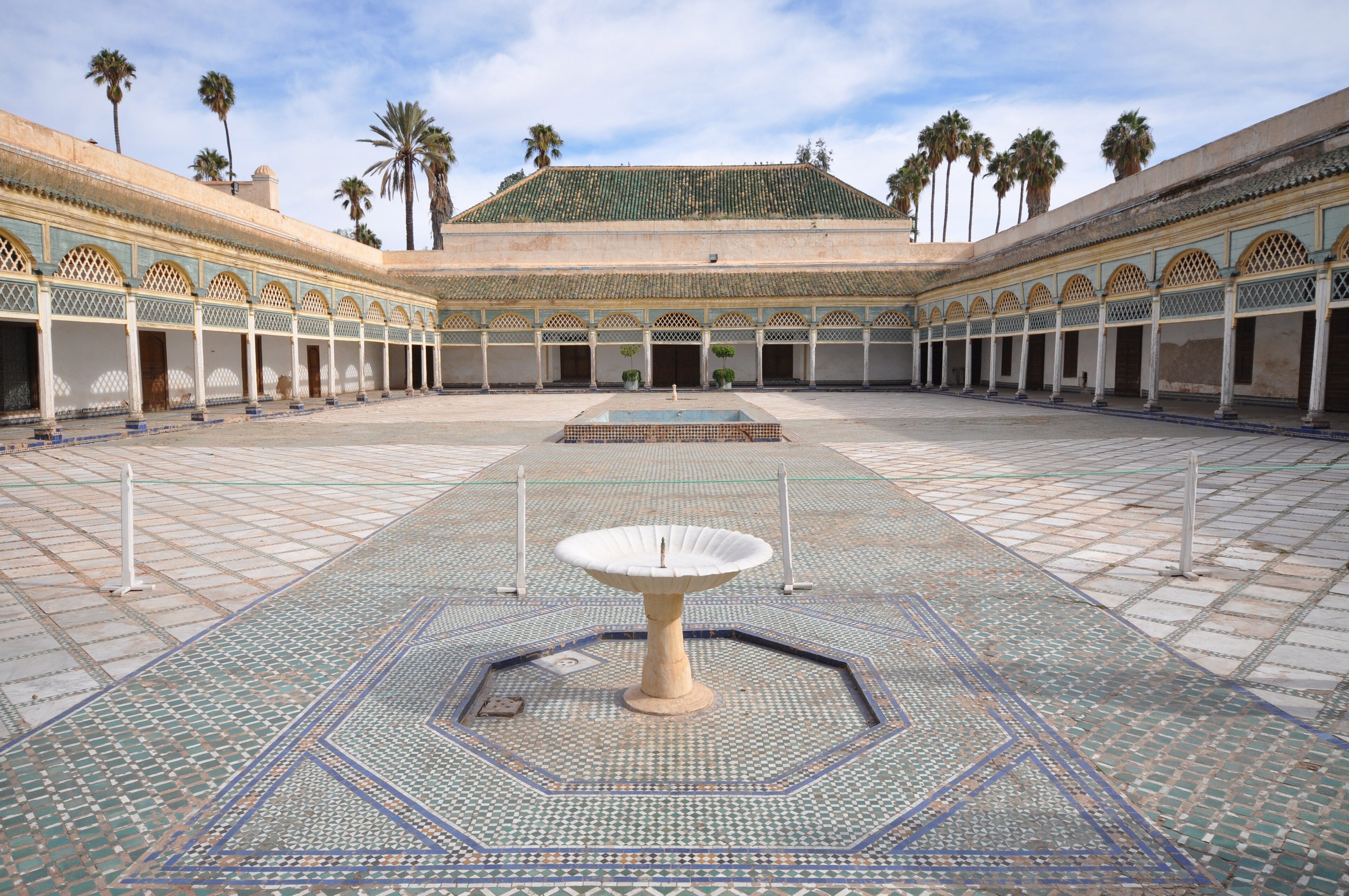 Il cortile del Palazzo Bahia a Marrakech, con fontana, mosaici azzurri e verdi, porticati decorati e palme sotto il cielo luminoso.